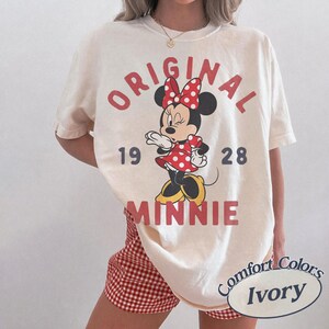 Op de afbeelding: Een ivoorkleurig T-shirt met korte mouwen en een afbeelding van Minnie Mouse in een rood-witte jurk met stippen en een strik. De tekst luidt ORIGINAL MINNIE 19 28 in rode en donkergrijze letters. Het shirt wordt gedragen met rood-witte ruitjesbroek.