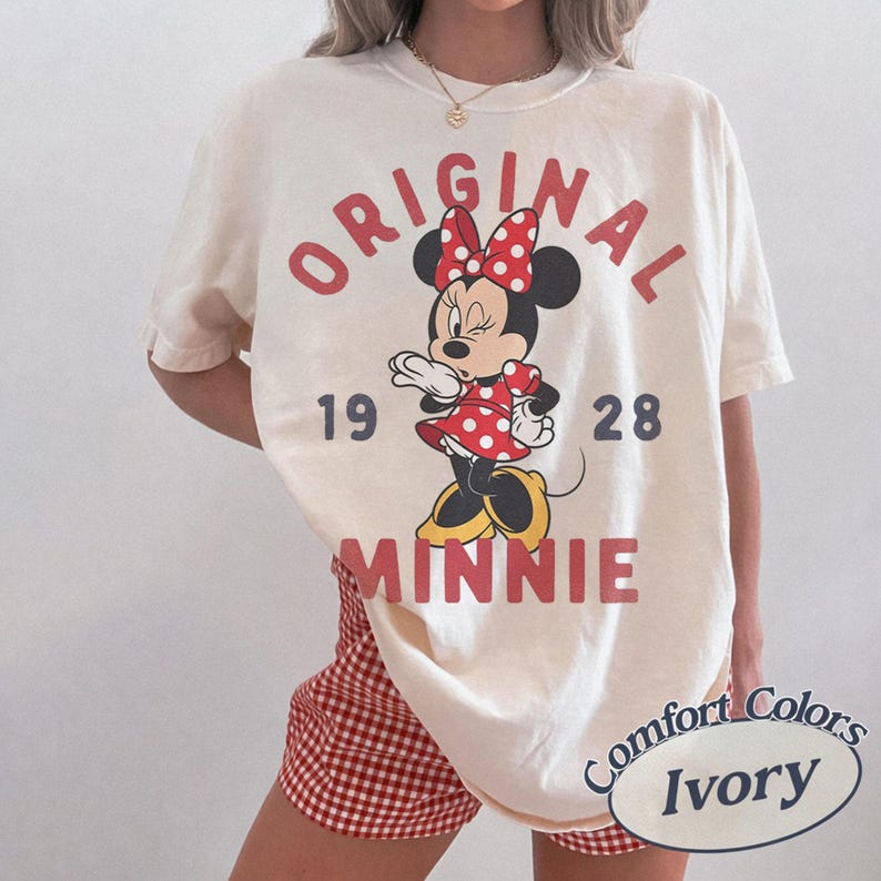 Op de afbeelding: Een ivoorkleurig T-shirt met korte mouwen en een afbeelding van Minnie Mouse in een rood-witte jurk met stippen en een strik. De tekst luidt ORIGINAL MINNIE 19 28 in rode en donkergrijze letters. Het shirt wordt gedragen met rood-witte ruitjesbroek.