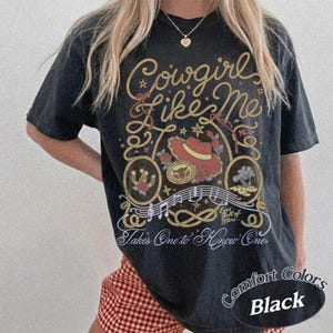 Op de afbeelding: Een zwart t-shirt met korte mouwen en een grafische print met de tekst Cowgirl like Me en Takes One to Know One. Het ontwerp bevat een touwrand, een rode cowboyhoed, een hoefijzer, muzieknoten en de tekst Eyes full of Stars.