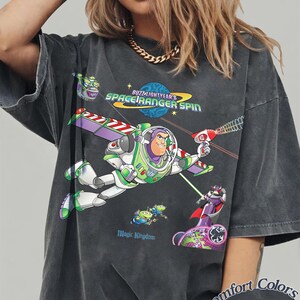 May include: Dark gray t-shirt featuring Buzz Lightyear from Toy Story. The graphic includes the text "Buzz Lightyear's Space Ranger Spin" and "Magic Kingdom". The shirt is a Comfort Colors Pepper tee.