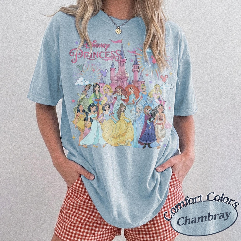 May include: Light blue t-shirt featuring a colorful graphic of Disney Princesses in front of a castle. The text "Disney Princess" is above the image. The shirt is a Comfort Colors Chambray.