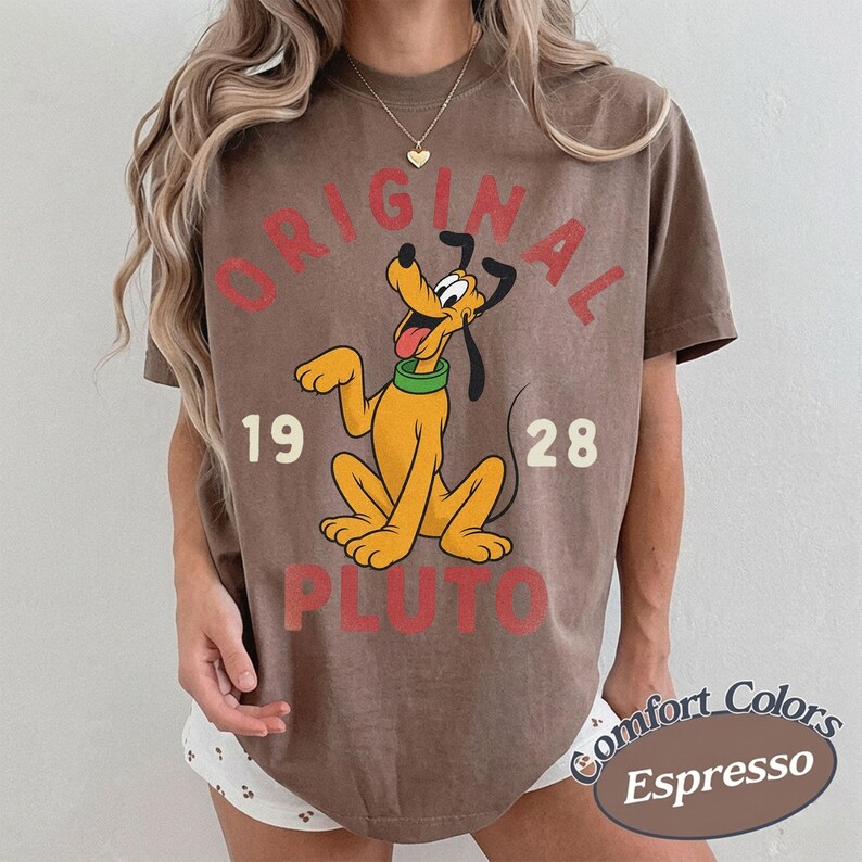 Op de afbeelding: Een bruin t-shirt met korte mouwen en een afbeelding van het Disney-personage Pluto. De tekst luidt ORIGINAL PLUTO met de cijfers 19 en 28 aan weerszijden van het personage. Het ontwerp is in rood en cr&egrave;me op de voorkant van het shirt gedrukt.