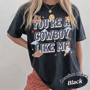 Op de afbeelding: Een zwart T-shirt met korte mouwen en de tekst YOU'RE A COWBOY LIKE ME in blauwe letters. Het ontwerp bevat illustraties van bruine cowboylaarzen, een bruine hoed, een gele ster, een raket, een bal en kleine rode en gele sterren.