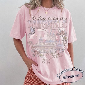 Może przedstawiać: Jasnor&oacute;żowy T-shirt z kr&oacute;tkim rękawem z nadrukiem graficznym przedstawiającym zamek, motyle i kwiaty. Tekst brzmi Today was a FAIRY TALE oraz Can you feel this magic in the air? Wz&oacute;r zawiera ilustracje liniowe w stonowanych odcieniach kolorystycznych.