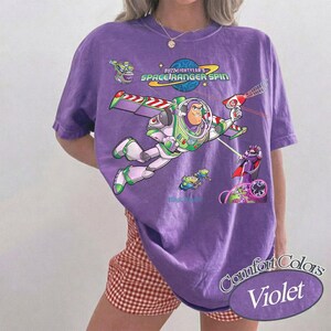 May include: A violet Comfort Colors t-shirt featuring Buzz Lightyear from Toy Story. The graphic includes Buzz in flight, the words "Buzz Lightyear's Space Ranger Spin," and other characters. The shirt has a relaxed fit.