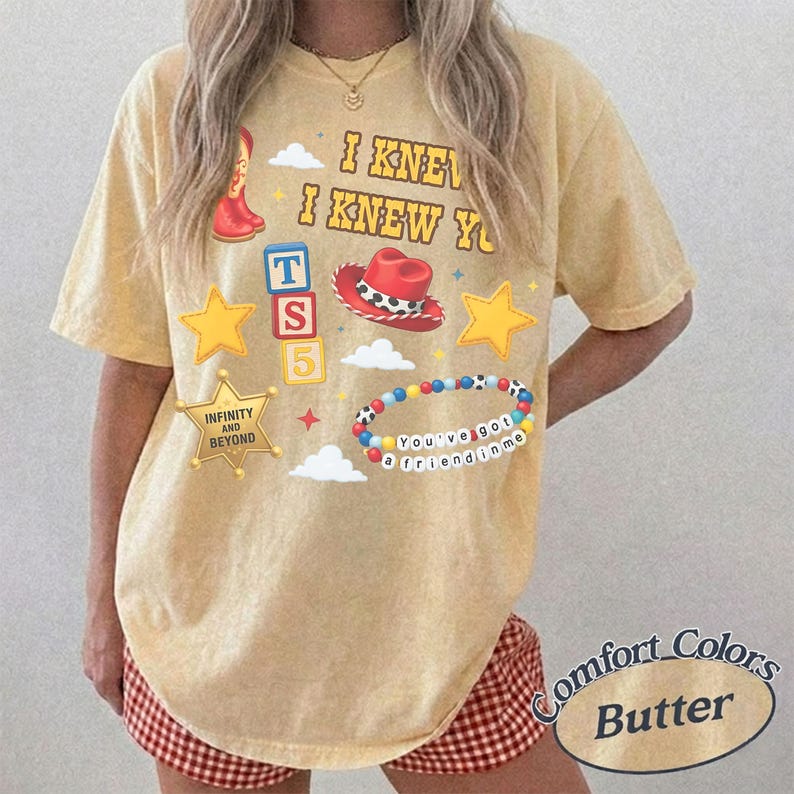 Pode incluir: Uma t-shirt amarela de manga curta com uma estampa gr&aacute;fica que inclui o texto I KNEW I KNEW YOU, uma bota de cowboy vermelha, um chap&eacute;u de cowboy vermelho, um bloco azul com TS5, estrelas amarelas e uma pulseira de contas coloridas.