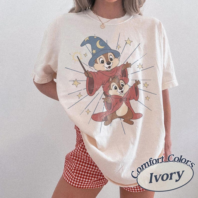 Op de afbeelding: Ivoor kleurig T-shirt met een cartoon illustratie van twee eekhoorns verkleed als tovenaars, met een toverstaf en sterren. Het shirt heeft de woorden "Comfort Colors" en "Ivory" erop gedrukt.