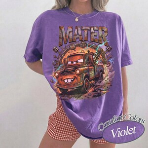 May include: A purple short-sleeve t-shirt featuring a graphic print of a rusty brown tow truck character with the text MATER above it. The design includes mechanical parts like pistons and wrenches surrounding the truck with lightning accents.
