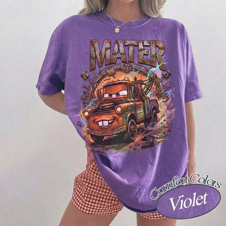 May include: A purple short-sleeve t-shirt featuring a graphic print of a rusty brown tow truck character with the text MATER above it. The design includes mechanical parts like pistons and wrenches surrounding the truck with lightning accents.