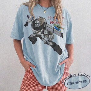 Pu&ograve; includere: T-shirt azzurra con una grafica di The Mandalorian e Grogu di Star Wars. Il design include il testo "Star Wars" in rosso. La maglietta &egrave; un Comfort Colors Chambray.