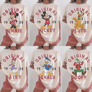 Op de afbeelding: Een collectie van zes cr&egrave;mekleurige T-shirts met Disney-figuren waaronder Minnie Mouse, Mickey Mouse, Pluto, Katrien Duck, Donald Duck en Goofy. Elk shirt toont de tekst ORIGINAL 1928 en de naam van het personage in het rood.