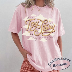Może przedstawiać: Jasnor&oacute;żowy T-shirt z kr&oacute;tkim rękawem z grafiką przedstawiającą ż&oacute;łtą linę ułożoną w napis "Toy Story" na tle białych chmur. Poniżej tekstu znajduje się napis "it's a" oraz "Baby, just say "yes"" wydrukowany ciemnobrązową czcionką pisankową.