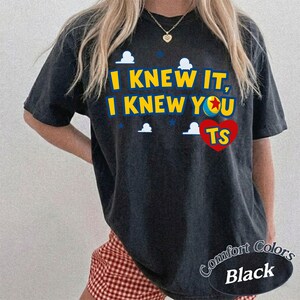 K&ouml;nnte beinhalten: Ein schwarzes T-Shirt mit kurzen &Auml;rmeln, das den gelben Text I KNEW IT, I KNEW YOU, eine bunte Ballgrafik anstelle des Buchstabens O, ein rotes Herz mit den Buchstaben TS und kleine wei&szlig;e Wolkensymbole auf der Vorderseite zeigt.