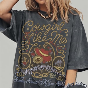 Op de afbeelding: Een antracietgrijs T-shirt met korte mouwen en een grafische print met de tekst Cowgirl Like Me en Takes One to Know One. Het ontwerp bevat een rode cowboyhoed, een lasso, muzieknoten, rozen en de zin Eyes full of Stars.