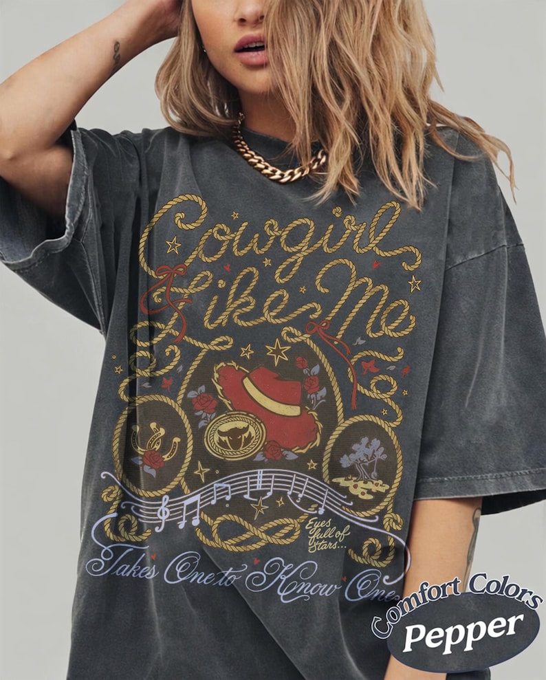 Op de afbeelding: Een antracietgrijs T-shirt met korte mouwen en een grafische print met de tekst Cowgirl Like Me en Takes One to Know One. Het ontwerp bevat een rode cowboyhoed, een lasso, muzieknoten, rozen en de zin Eyes full of Stars.