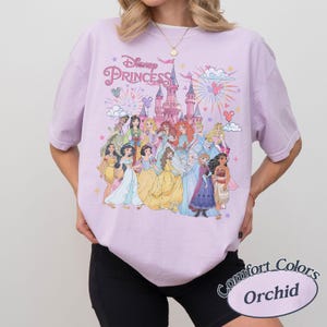 May include: A light purple t-shirt featuring a graphic of Disney Princesses in front of a castle with fireworks. The text "Disney Princess" is above the graphic. The shirt is a Comfort Colors Orchid.