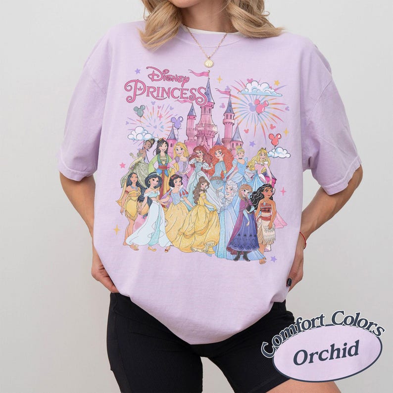 May include: A light purple t-shirt featuring a graphic of Disney Princesses in front of a castle with fireworks. The text "Disney Princess" is above the graphic. The shirt is a Comfort Colors Orchid.