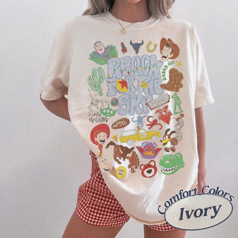 Peut inclure: Un t-shirt ivoire avec une impression graphique color&eacute;e portant le texte "REACH FOR THE SKY" et divers personnages de dessins anim&eacute;s et ic&ocirc;nes, dont un cow-boy, un ranger de l'espace, un dinosaure, un extraterrestre et un camion jaune.