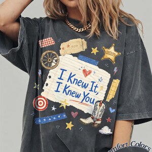 Puede incluir: Una camiseta gris carb&oacute;n con un gr&aacute;fico central de una p&aacute;gina de cuaderno rasgada con el texto "I Knew It, I Knew You" en azul. El dise&ntilde;o incluye una bota de vaquero, una estrella de sheriff, una rueda de carreta y peque&ntilde;os iconos.