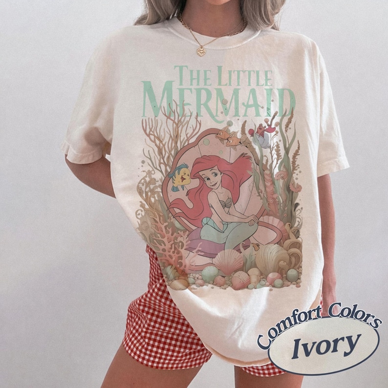 Pu&ograve; includere: Una t-shirt a maniche corte color avorio con una stampa grafica de La Sirenetta che raffigura Ariel, Flounder ed elementi di corallo sottomarino. Il design include il testo The Little Mermaid in un carattere verde chiaro. Girocollo.