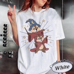 Op de afbeelding: Wit T-shirt met een cartoon illustratie van twee chipmunks in tovenaarskostuums, compleet met hoeden en capes. Het ontwerp bevat sterren en een toverstaf. De tekst "Comfort Colors White" staat rechtsonder.