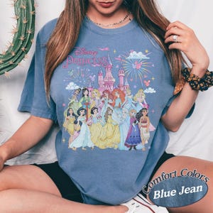May include: Blue jean Comfort Colors t-shirt featuring a graphic of Disney Princesses in front of a castle with fireworks. The text "Disney Princess" is above the castle. The shirt is short-sleeved.