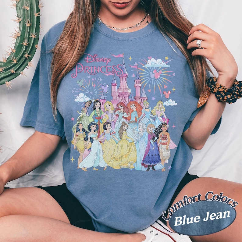 May include: Blue jean Comfort Colors t-shirt featuring a graphic of Disney Princesses in front of a castle with fireworks. The text "Disney Princess" is above the castle. The shirt is short-sleeved.