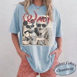May include: Light blue t-shirt featuring a vintage graphic of Mickey Mouse and Walt Disney. The graphic is in black and white with the word "WALT" in red. The shirt is a Comfort Colors Chambray.