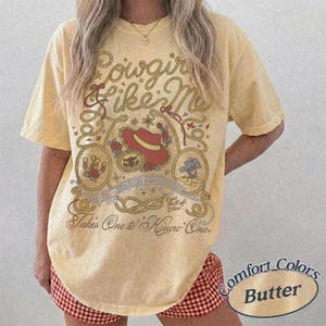 Op de afbeelding: Een lichtgeel t-shirt met korte mouwen en een bruin touwontwerp, een rode cowboyhoed, een hoefijzer en muzieknoten. De tekst luidt Cowgirl Like Me en Takes One to Know One, inclusief de zin Eyes full of Stars.