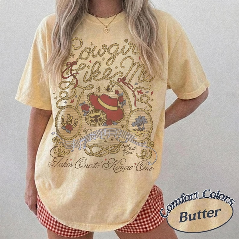 Op de afbeelding: Een lichtgeel t-shirt met korte mouwen en een bruin touwontwerp, een rode cowboyhoed, een hoefijzer en muzieknoten. De tekst luidt Cowgirl Like Me en Takes One to Know One, inclusief de zin Eyes full of Stars.