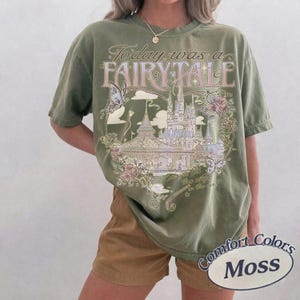 Może przedstawiać: Oliwkowozielony t-shirt z kr&oacute;tkim rękawem z nadrukiem graficznym przedstawiającym zamek, motyle i kwiaty. Tekst brzmi Today was a FAIRY TALE. Wz&oacute;r zawiera ozdobne zawijasy i gwiazdki w odcieniach r&oacute;żu, błękitu i kremu.