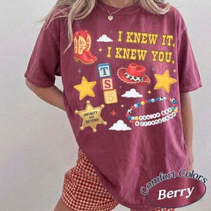 Pode incluir: Uma t-shirt de manga curta cor de vinho com uma estampa gr&aacute;fica que inclui o texto I KNEW IT, I KNEW YOU, uma bota de cowboy vermelha, um chap&eacute;u de cowboy vermelho, estrelas amarelas, um distintivo de xerife com INFINITY AND BEYOND e blocos TS 5.