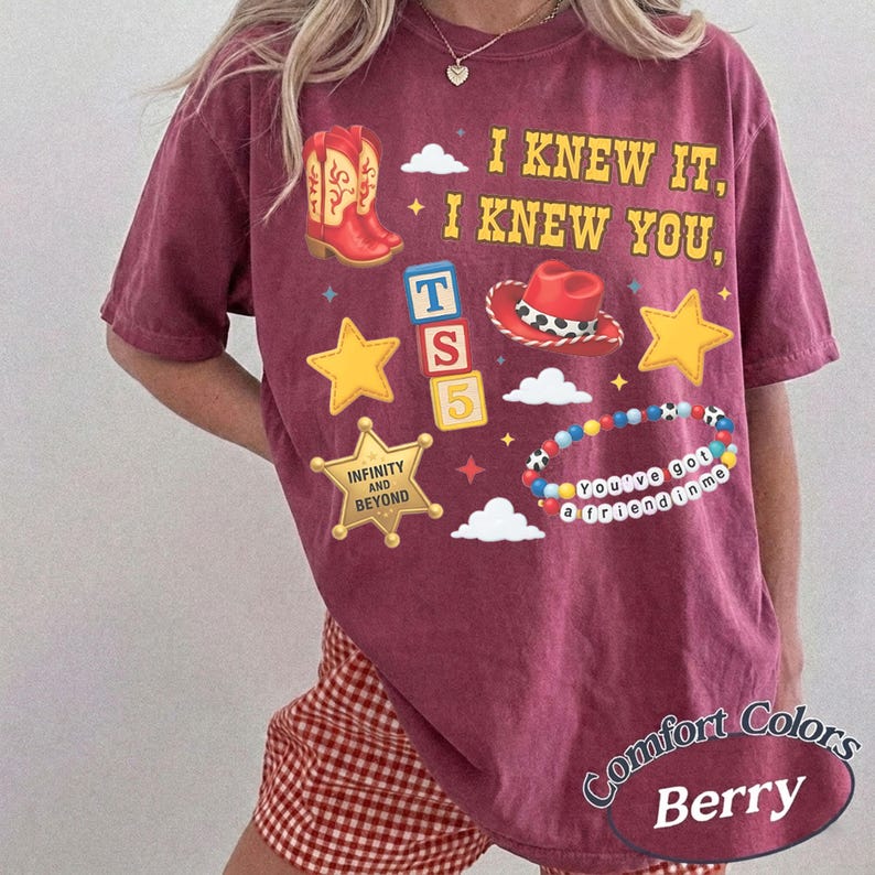 Pode incluir: Uma t-shirt de manga curta cor de vinho com uma estampa gr&aacute;fica que inclui o texto I KNEW IT, I KNEW YOU, uma bota de cowboy vermelha, um chap&eacute;u de cowboy vermelho, estrelas amarelas, um distintivo de xerife com INFINITY AND BEYOND e blocos TS 5.
