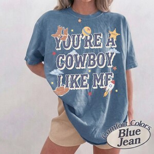 Op de afbeelding: Een blauw T-shirt met korte mouwen en de tekst "YOU'RE A COWBOY LIKE ME" in het wit met een donkere omlijning. De afbeelding bevat illustraties van bruine cowboylaarzen, een gele bal, een sheriffster, een bruine hoed en een raket.