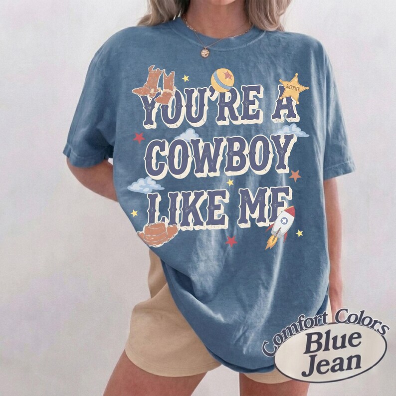 Op de afbeelding: Een blauw T-shirt met korte mouwen en de tekst "YOU'RE A COWBOY LIKE ME" in het wit met een donkere omlijning. De afbeelding bevat illustraties van bruine cowboylaarzen, een gele bal, een sheriffster, een bruine hoed en een raket.