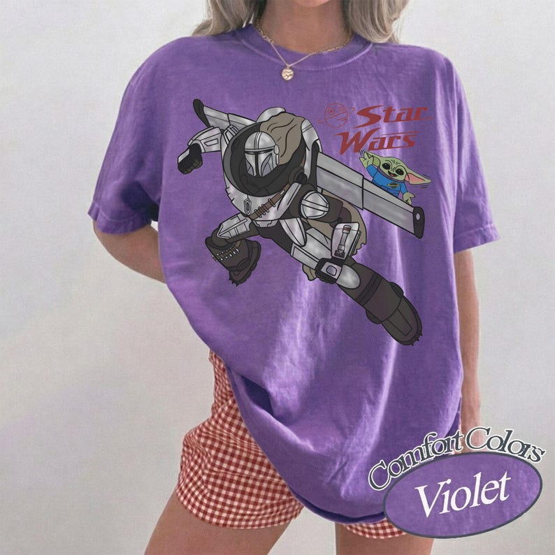 Pu&ograve; includere: T-shirt Comfort Colors viola con una grafica di The Mandalorian e Grogu di Star Wars. Il design include il testo "Star Wars" in rosso. La maglietta &egrave; di colore viola unito.