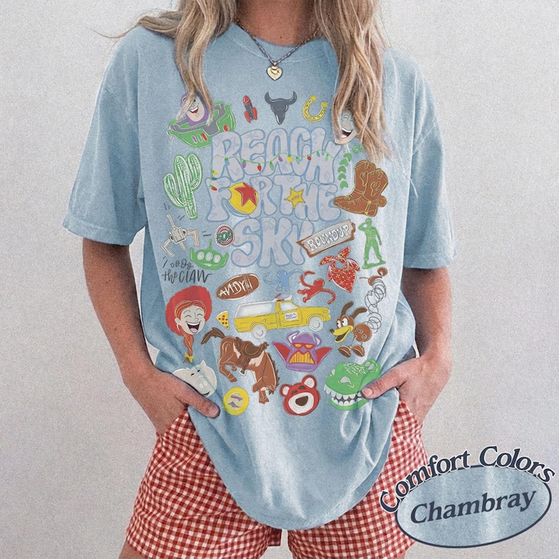 Peut inclure: Un t-shirt bleu clair &agrave; manches courtes avec une impression graphique portant le texte Reach for the Sky et divers personnages et ic&ocirc;nes de dessins anim&eacute;s color&eacute;s, dont un chapeau de cow-boy, un camion jaune, un extraterrestre vert.