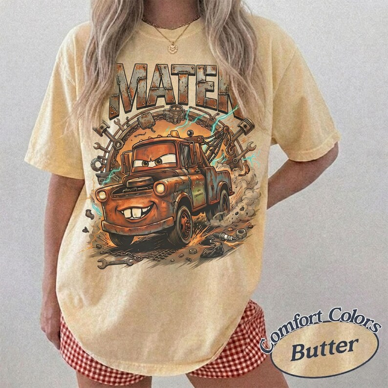 May include: A butter-yellow short-sleeve t-shirt featuring a graphic print of a rusty brown tow truck character with the text MATER above it. The design includes mechanical tools like wrenches and gears surrounding the truck with dust clouds.