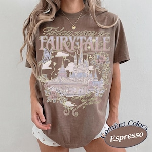 Może przedstawiać: Brązowy t-shirt z kr&oacute;tkim rękawem z nadrukiem graficznym przedstawiającym zamek, motyle i kwiaty. Tekst brzmi Today was a FAIRY TALE oraz Can you feel the magic in the air? Wz&oacute;r zawiera jasnoniebieskie, r&oacute;żowe i kremowe akcenty kolorystyczne.