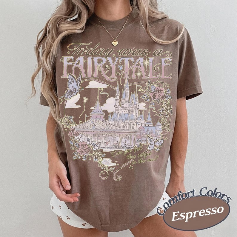 Może przedstawiać: Brązowy t-shirt z kr&oacute;tkim rękawem z nadrukiem graficznym przedstawiającym zamek, motyle i kwiaty. Tekst brzmi Today was a FAIRY TALE oraz Can you feel the magic in the air? Wz&oacute;r zawiera jasnoniebieskie, r&oacute;żowe i kremowe akcenty kolorystyczne.