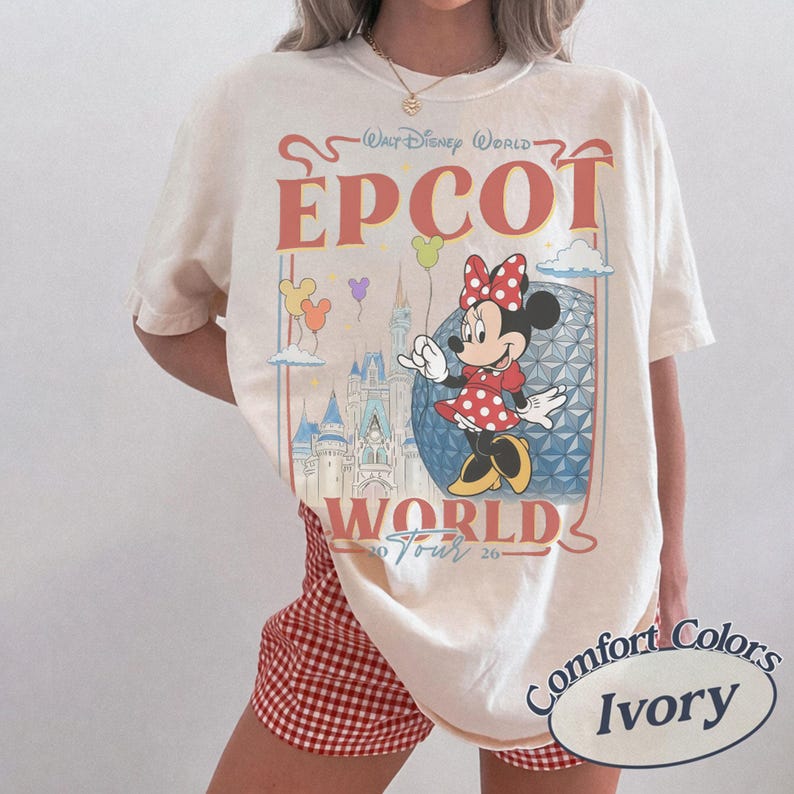以下が含まれることがあります： アイボリーのTシャツで、ミニーマウス、エプコットのジオデシック・スフィア、城のグラフィックプリントが施されています。テキストには「Walt Disney World」、「EPCOT」、「World Tour 2026」と書かれています。赤、青、黄色のアクセントと浮かぶ風船がデザインされています。