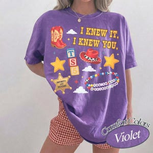 Pode incluir: Uma t-shirt roxa de manga curta com uma estampa gr&aacute;fica que inclui o texto I KNEW IT, I KNEW YOU, uma bota de cowboy vermelha, um chap&eacute;u de cowboy vermelho, blocos empilhados com as letras TS 5, uma estrela dourada e uma pulseira de contas.