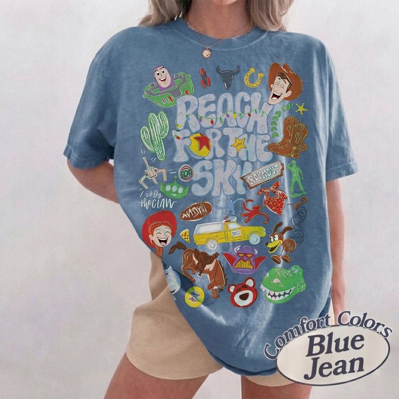 Peut inclure: Un t-shirt bleu &agrave; manches courtes avec une impression graphique portant le texte REACH FOR THE SKY et divers personnages et ic&ocirc;nes de dessins anim&eacute;s, notamment un cow-boy, un ranger de l'espace, un camion jaune, un cactus et un dinosaure vert.