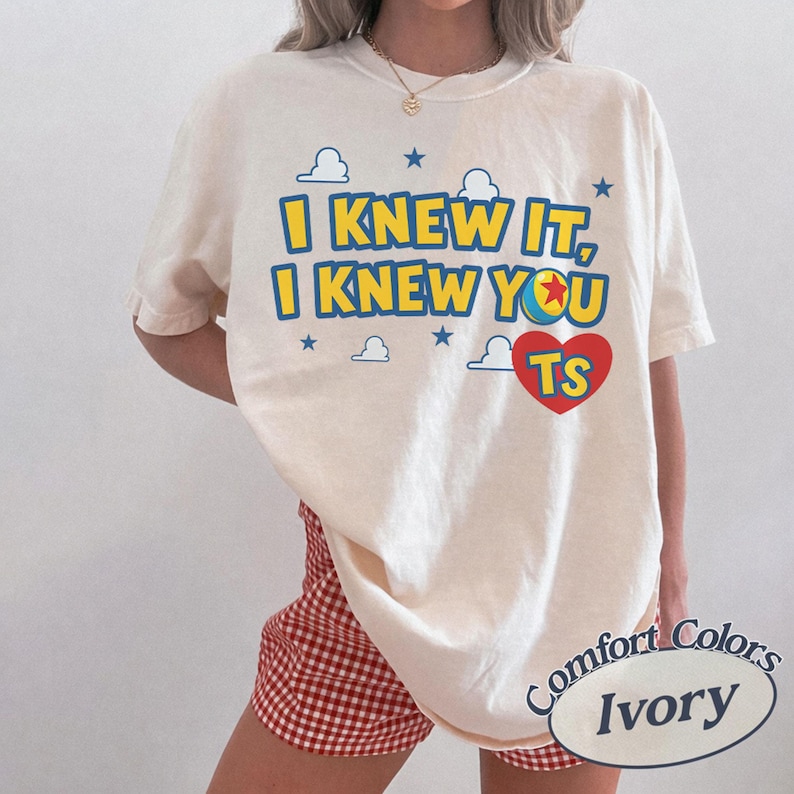 K&ouml;nnte beinhalten: Ein elfenbeinfarbenes Kurzarm-T-Shirt mit dem blau-gelben Text I KNEW IT, I KNEW YOU und einem roten Herz mit den Buchstaben TS. Das Design enth&auml;lt kleine blaue Sterne und wei&szlig;e Wolkenumrisse, wobei eine Ballgrafik den Buchstaben O ersetzt.