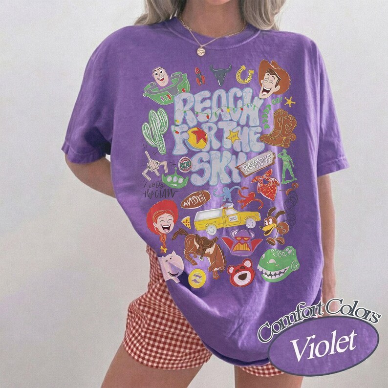 Peut inclure: Un t-shirt violet &agrave; manches courtes avec une impression graphique portant le texte Reach For The Sky et diverses illustrations de personnages de dessins anim&eacute;s, dont un cow-boy, un ranger de l'espace, un dinosaure, un cochon, un ours et un camion jaune.