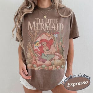 Pu&ograve; includere: Una t-shirt marrone a maniche corte con una stampa grafica de La Sirenetta che raffigura Ariel seduta in una conchiglia circondata da coralli e vita marina. Il testo The Little Mermaid &egrave; stampato in un carattere serif sopra l'illustrazione.