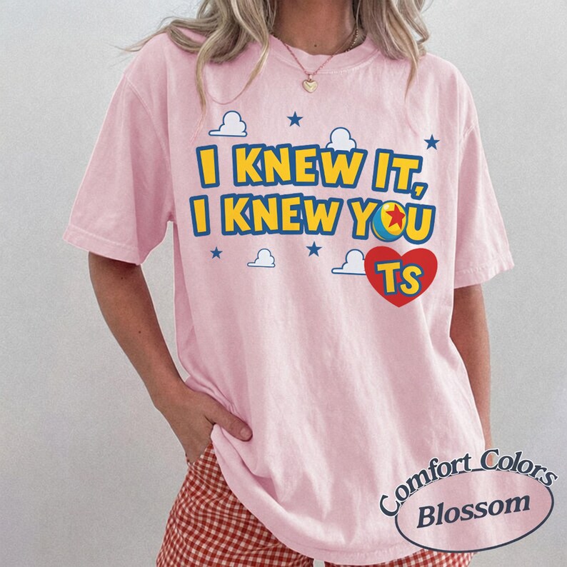 K&ouml;nnte beinhalten: Ein hellrosa T-Shirt mit kurzen &Auml;rmeln und dem Text I KNEW IT, I KNEW YOU TS in gelben und blauen Buchstaben. Das Design enth&auml;lt einen bunten Ball, kleine blaue Sterne, wei&szlig;e Wolkenumrisse und ein rotes Herz mit den Buchstaben TS.