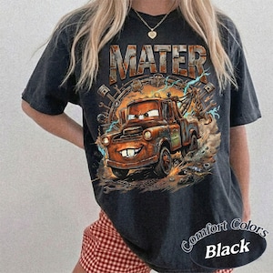 May include: A black short-sleeve t-shirt featuring a graphic print of a rusty brown tow truck character with large eyes and buck teeth. The text MATER is printed in a metallic font above the truck, surrounded by mechanical parts and lightning.