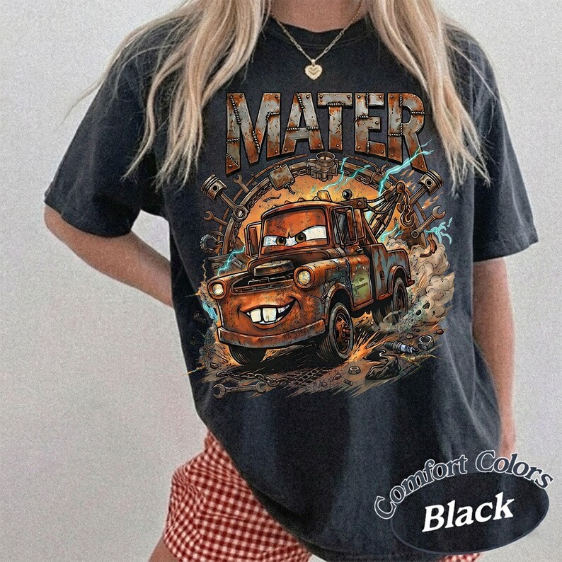 May include: A black short-sleeve t-shirt featuring a graphic print of a rusty brown tow truck character with large eyes and buck teeth. The text MATER is printed in a metallic font above the truck, surrounded by mechanical parts and lightning.