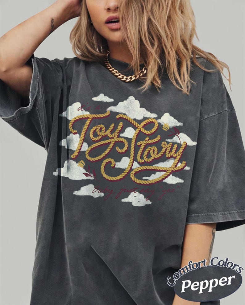 Może przedstawiać: Ciemnoszary t-shirt oversize z grafiką białych chmur i napisem Toy Story wykonanym ż&oacute;łtą czcionką przypominającą linę. Wz&oacute;r zawiera małe czerwone kwiatowe akcenty oraz napis baby just say yes wykonany cienką, czerwoną czcionką pisankową.